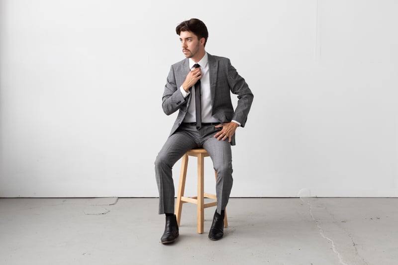 Indochino Harrogate Glen Check Suit straightening tie while sitting on stool