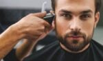 Bearded man looking directly at the camera and getting his sideburns tapered with clippers